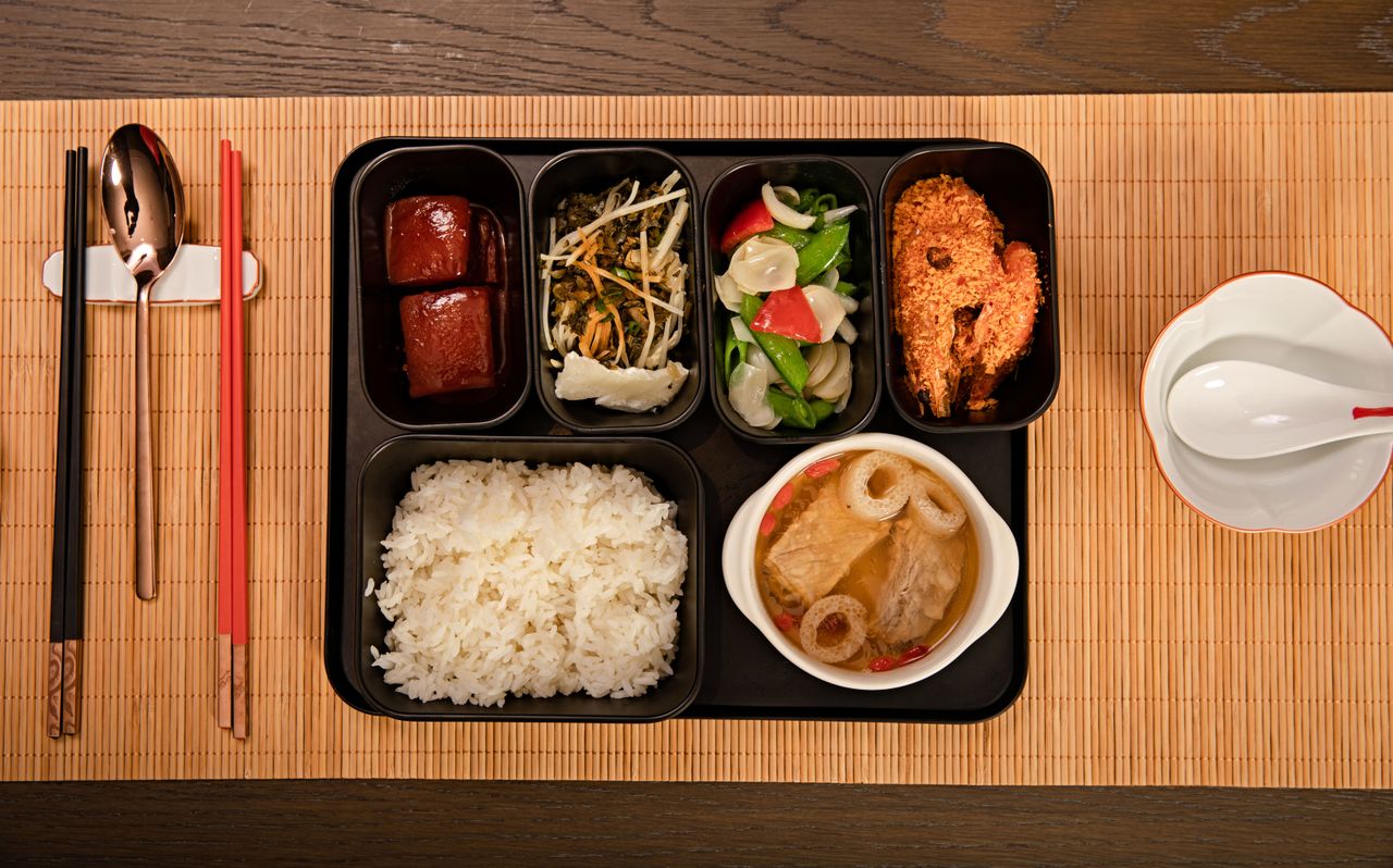 Bento tray with individual food compartments alongside a ceramic soup bowl, metal spoon and chopsticks, showing a mix of materials used around a bento meal