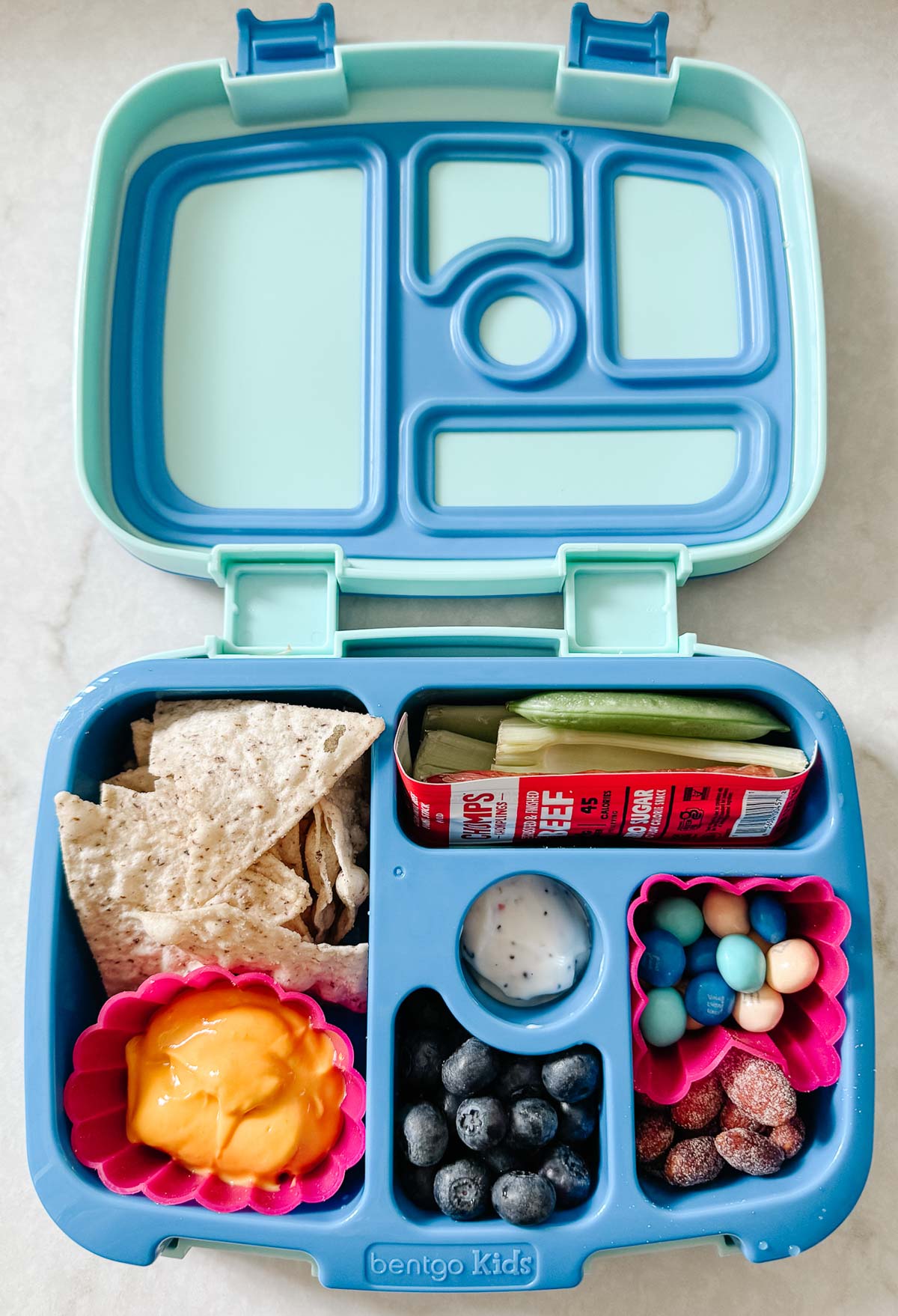 Open kids bento box neatly packed with crackers, cheese, vegetables, a beef stick, yogurt dip, blueberries and a small treat cup in each compartment